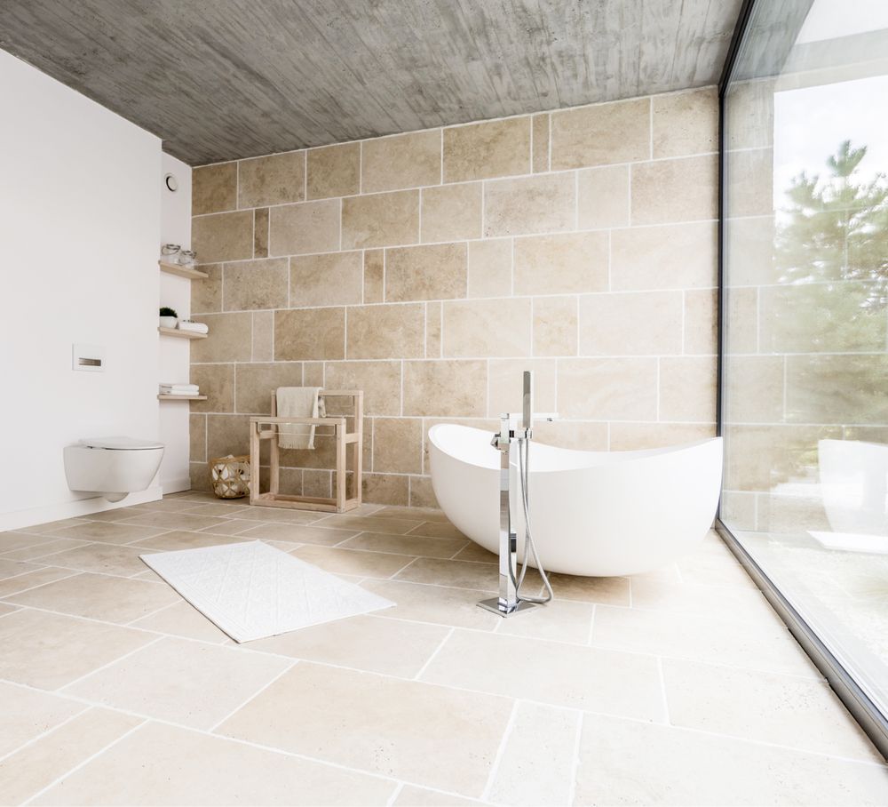 Modernes Badezimmer mit freistehender weißer Badewanne, Wänden aus Steinfliesen, einem Holzstuhl und einem großen Glasfenster für natürliches Licht.