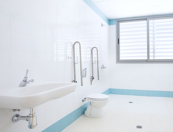 Ein helles, modernes Badezimmer mit weißem Waschbecken, Toilette, Haltegriffen und einem Fenster mit horizontalen Jalousien. Klares und minimalistisches Design.