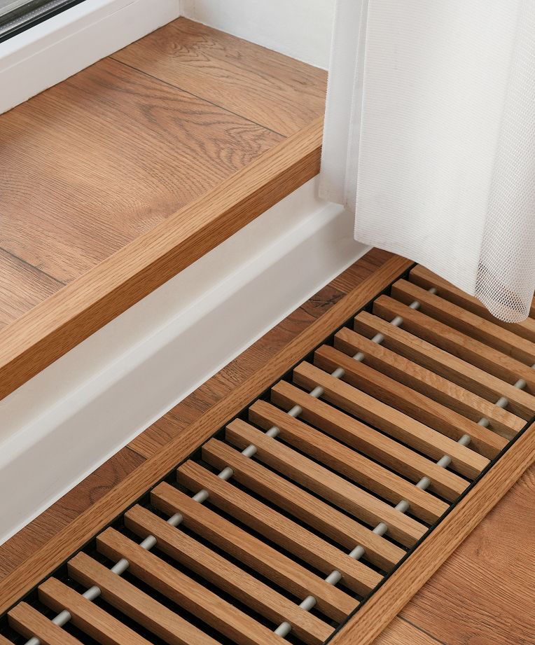 Nahaufnahme eines Holzbodenübergangs mit einer Lamellenheizung unter einem weißen Vorhang, der warme Töne und modernes Design zur Schau stellt.